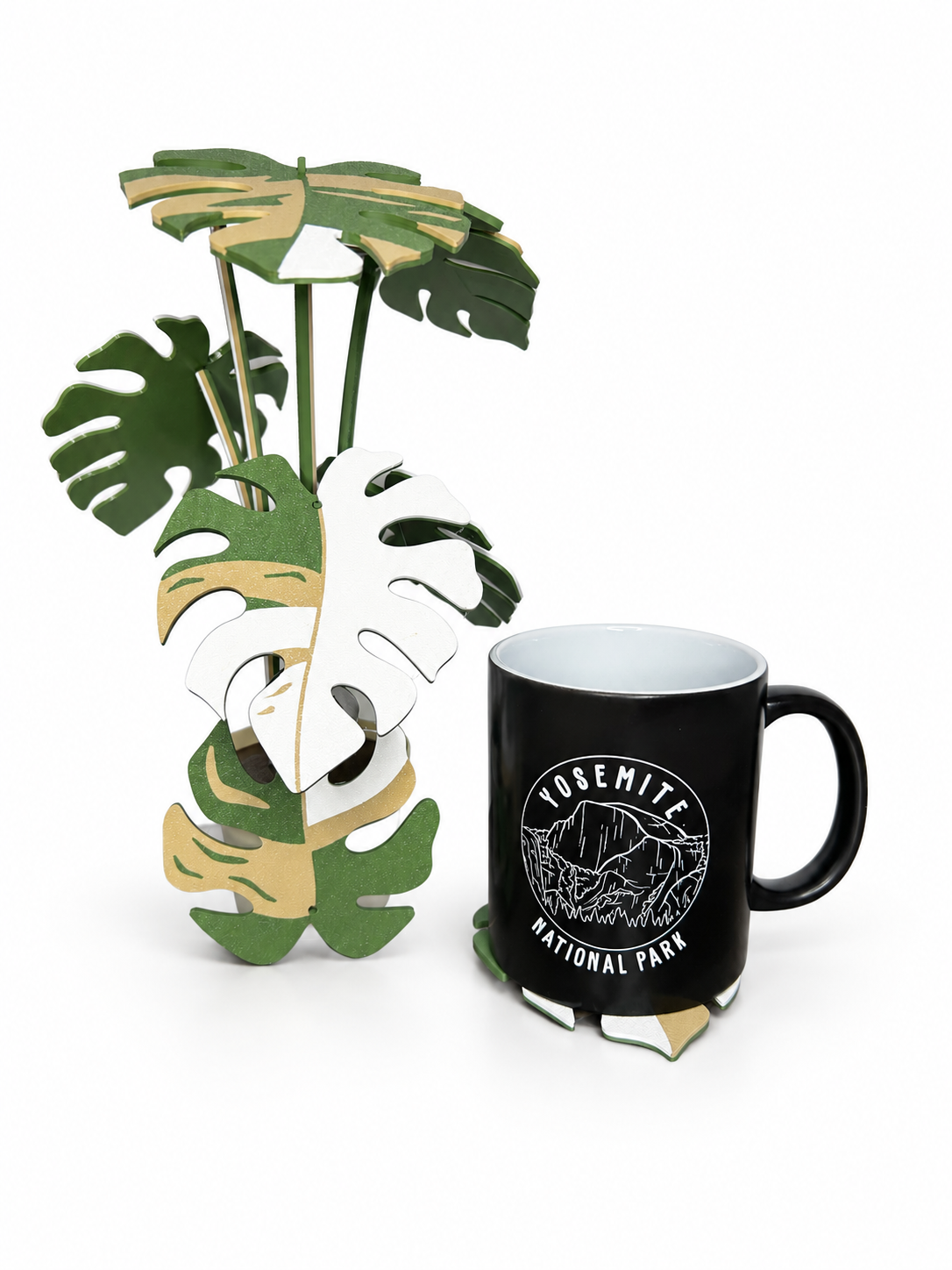 Beige, white and green Monstera coaster display beside a mug on a leaf coaster, photographed on a white background.